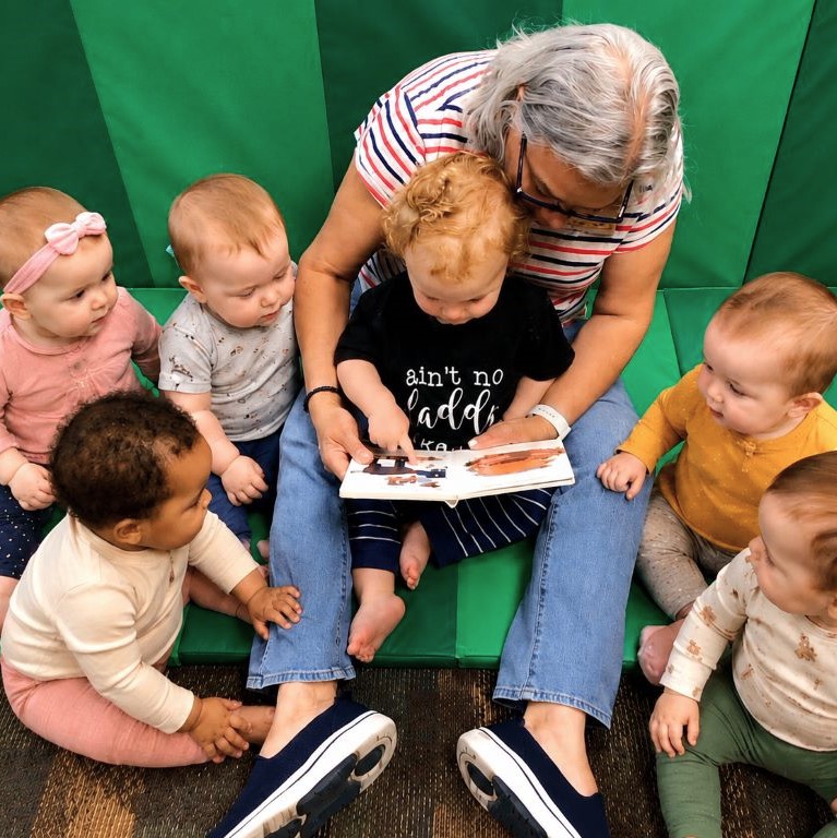 Edina Daycare teacher reading a book to a class of infants at Edina Daycare Spanish Immersion, with one child on the adult’s lap pointing at the pages.